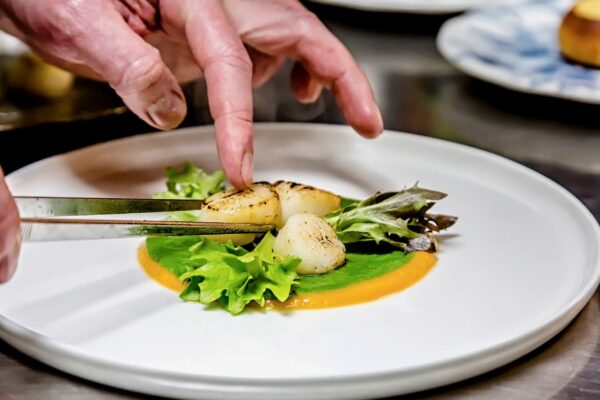 Echilibrul culorilor Chef Silviu aranjează medalioane de scoici pe un sos verde și portocaliu, completat cu frunze proaspete.