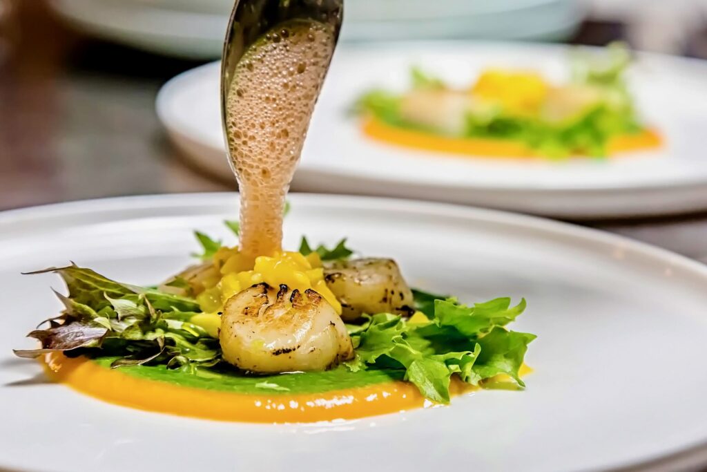 Respirația gustului Chef Silviu adaugă o spumă aerată peste scoici caramelizate, servite cu sos verde, portocaliu și frunze proaspete